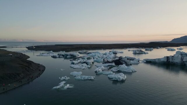 Golden hour aerial flight reflecting warm sunset light on blue ice chunks at J&ouml;kuls&aacute;rl&oacute;n lagoon, Iceland.