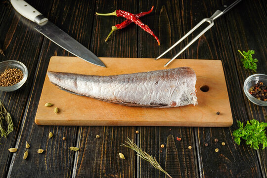 A Merluccius fish fillet is placed on a wooden cutting board. Fresh spices and cooking tools surround the fillet. The setting is a kitchen with a rustic look