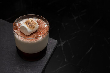 A close-up shot of an elegant cocktail featuring a marshmallow. The drink is served in a stylish glass on a dark surface, creating a sophisticated ambiance