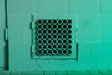 A small square metal ventilation grille with round tube pattern on a painted tiled wall, close-up, mild wear and dust.