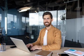 Businessman with a beard, wearing an orange shirt, working on a laptop at his desk, smiling at the...