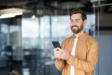 Smiling man standing in a modern office space, actively engaging with his smartphone, showcasing...