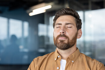 Man with closed eyes stands in a modern office, practicing deep breathing and meditation for stress relief, mental health, and employee wellbeing during a work break