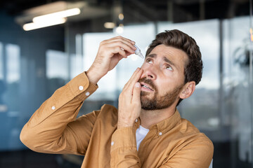 Man holding an eye dropper, tilting his head back to apply drops into his eye, while touching his...