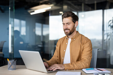 Young smiling businessman in casual shirt working remotely on a laptop in a contemporary office setting, engaging in a video call or listening to a podcast using wireless earbuds