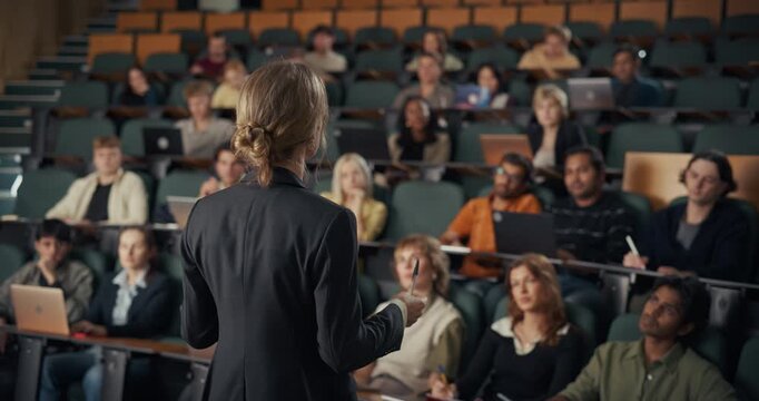 Middle‑aged White Female Professor Delivers University Lecture, Gesturing With a Pen While a Diverse Group of College Students With Laptops,  Active Learning Inside a Large Modern Academic Auditorium.