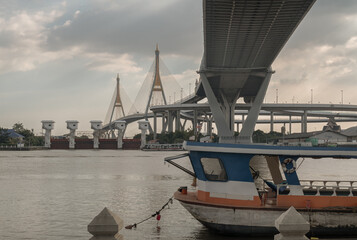 The majestic cable-stayed Bhumibol Bridge crosses the Chao Phraya River in Bangkok with a small boat moored prominently in the foreground.
