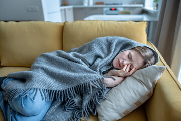 Sick woman wrapped in blanket lying on sofa at home, experiencing chills, body aches, fatigue. Female feeling unwell during cold winter season touching face with closed eyes suffering headache