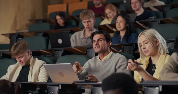 In University Lecture Hall, a Male Student Surrounded by Diverse Classmates Listens Intently, Raises His Hand, and Confidently Answers the Professor&rsquo;s Question, Using a Laptop