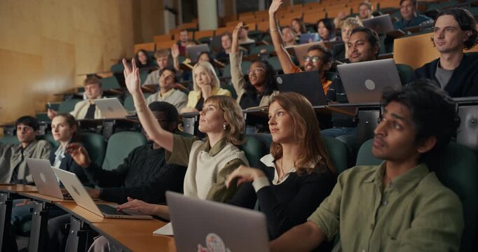 University Students Sit in a Modern Lecture Hall With Open Laptops, Listening to Instructor, Taking Notes, and Raising Hands to Contribute Questions and Answers During an Interactive Higher Education
