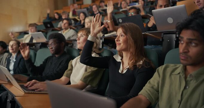 Engaged University Students Sit in Lecture Hall With Open Laptops, Listening Attentively, Taking Notes, and Raising Hands to Ask Questions, Understanding Course Material and Critical Thinking.