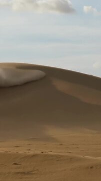 Wind blows dust off a sand dune in the desert under a blue sky during the day