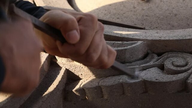 Close up of artisan hands carving stone with chisel and hammer. Skilled sculptor creating detailed architectural ornament. Traditional manual craftsmanship process in a workshop setting.

