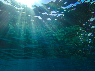 Obraz premium Dark blue ocean surface seen from underwater. Abstract waves underwater and rays of sunlight shining through, Sun light rays undersea deep, Underwater background with sea bottom, Mediterranean sea.