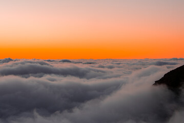 A sea of clouds rolls over mountain peaks at Pico do Arieiro, Madeira, Portugal. Warm orange to pink sky, dark rock slope at right, faint ridgelines and turbines.