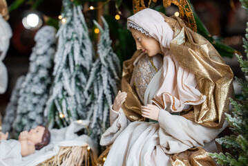 Mary in white and gold robes gazes toward infant Jesus in a straw crib. Snow dusted evergreens and...