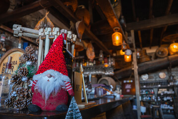 Warm pendant lights glow over a wooden counter with a Santa gnome and pinecones in Funchal, Madeira. Hams hang near wine barrels and bottles, evoking poncha culture in December.