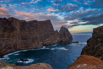Sheer volcanic cliffs at Ponta de Sao Lourenco, Madeira, Portugal, with waves foaming around jagged sea stacks under pastel clouds, seen from a high rocky vantage point.