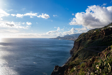Rugged cliffs of Madeira descend to the Atlantic, Funchal lines the crescent bay, late afternoon light reflects on calm water, clouds drift over terraced hillsides.