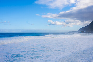 Powerful Atlantic swells crash onto a wide foamy shore, dark cliffs rise right, a small coastal settlement sits nearby in Madeira, Portugal, soft afternoon light glows.
