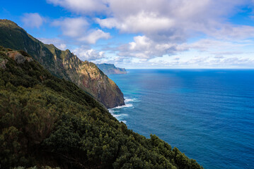 Steep cliffs on Madeira north coast meet the Atlantic under midday sun, with laurel like shrubs, turquoise water bands, and surf tracing bases toward Ponta de Sao Lourenco.