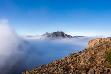 Jagged Pico do Arieiro rises above a sea of clouds in Madeira, Portugal, under warm morning light, with mist in valleys and wide angle view emphasizing elevation.