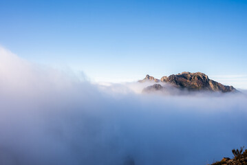Pico do Arieiro in Madeira, Portugal rises above a sea of clouds as warm sunlight grazes craggy rock faces, wide composition, soft early day light, weather inversion present
