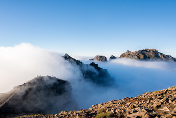 Rugged volcanic peaks at Pico do Arieiro in Madeira, Portugal rise above low clouds as hikers move along the path. Cool daylight highlights rock while mist swirls in valleys.