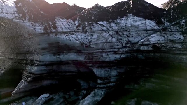 Cinematic aerial drone flight through Katla ice cave tunnel showing blue ice with black volcanic ash veins and green mossy mountains in Iceland 4K.