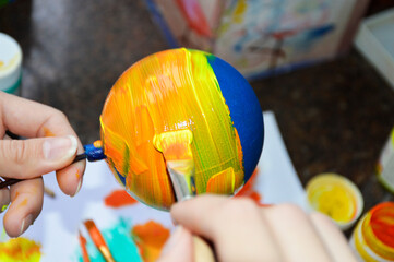Women's hands carefully paint a brightly colored Christmas ball: with a brush they draw stripes of paint on its surface - the basis for the drawing