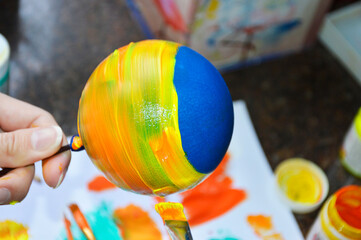 Women's hands carefully paint a brightly colored Christmas ball: with a brush they draw stripes of paint on its surface - the basis for the drawing