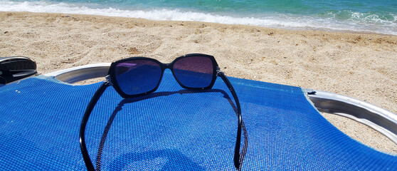 A pair of dark-framed sunglasses rests on a blue beach chair by the sea. A sandy beach and turquoise water with gentle waves are visible in the background. A sunny day, relaxation, and carefree mood.