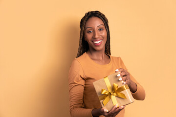 joyful african american woman with gift box present in beige colors. christmas, happy birthday, celebration concept.