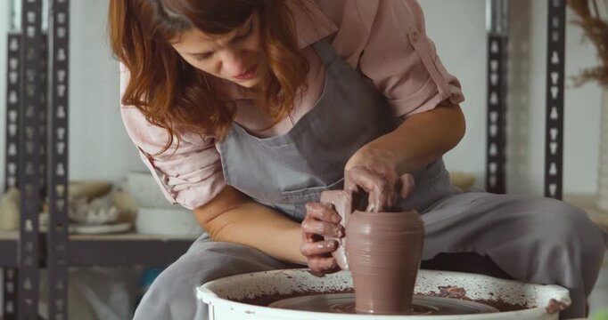 A woman in a pink shirt and apron shapes clay on a spinning wheel, carefully forming a pot with muddy hands in a ceramics studio. Zoom in camera shot.