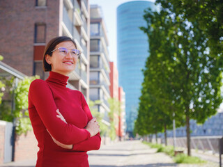Woman in glasses and red turtleneck, dressed in business style, stands in center of Frankfurt, surrounded by modern buildings and urban life, embodying professionalism and confidence