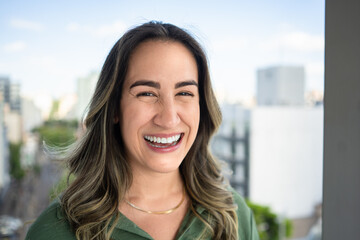 Joyful Latin businesswoman in her 30s laughing with genuine happiness wearing green shirt against blurred urban cityscape background