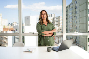 Latin businesswoman in her 30s with bright smile wearing green shirt standing confidently with arms crossed in modern office with cityscape view and laptop on white desk