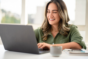 Cheerful Latin businesswoman in her 30s wearing olive green shirt working on laptop with genuine smile in bright modern office with coffee mug and notebook on white desk