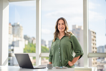 Confident Latin businesswoman standing beside desk with hands in pockets smiling in modern office with panoramic windows showcasing urban skyline and greenery while laptop and coffee mug