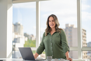 Confident Latin businesswoman in green button-up shirt smiling while standing at white desk with laptop and coffee mug in bright modern office 