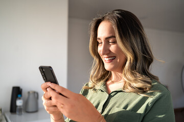 Smiling Latin businesswoman in green button-up shirt looking at smartphone with joyful expression in home office with natural sunlight creating dramatic lighting on her face