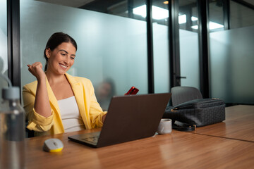 Excited professional woman in yellow blazer celebrating success while working on laptop in modern corporate office