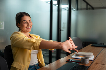 Happy professional woman in yellow blazer stretching arms and wrists during work break in modern...