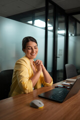 Happy businesswoman in yellow blazer enjoying remote work on laptop in contemporary corporate office environment