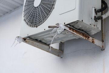 Photo of air conditioner split system outdoor unit in winter with some icicles built up. Outdoor unit of air conditioner in the wall of building under roof. Heating with air conditioner concept.