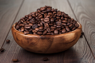 Close up photo of roasted coffee beans in bowl. Wooden bowl full of roasted coffee beans on wooden background with some beans scattered nearby.