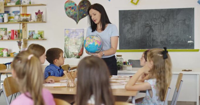 Teacher and elementary students learning geography with globe in classroom, curious children exploring world map together, social studies and global education for kids.