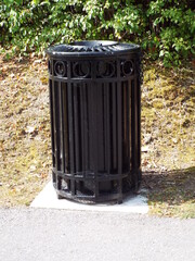 A cylindrical "black metal public trash can" with vertical slats and an ornate top ring, sitting on a square concrete base next to a paved path and a grassy bank.