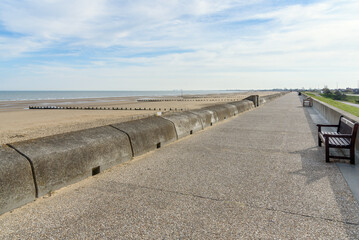 Deserted footpath lined with wooden benches on a seawall running along a large sandy beach on a partly cloudy spring day