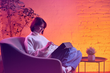 Woman using tablet with stylus in cozy chair under neon warm lighting. Concept of creative...